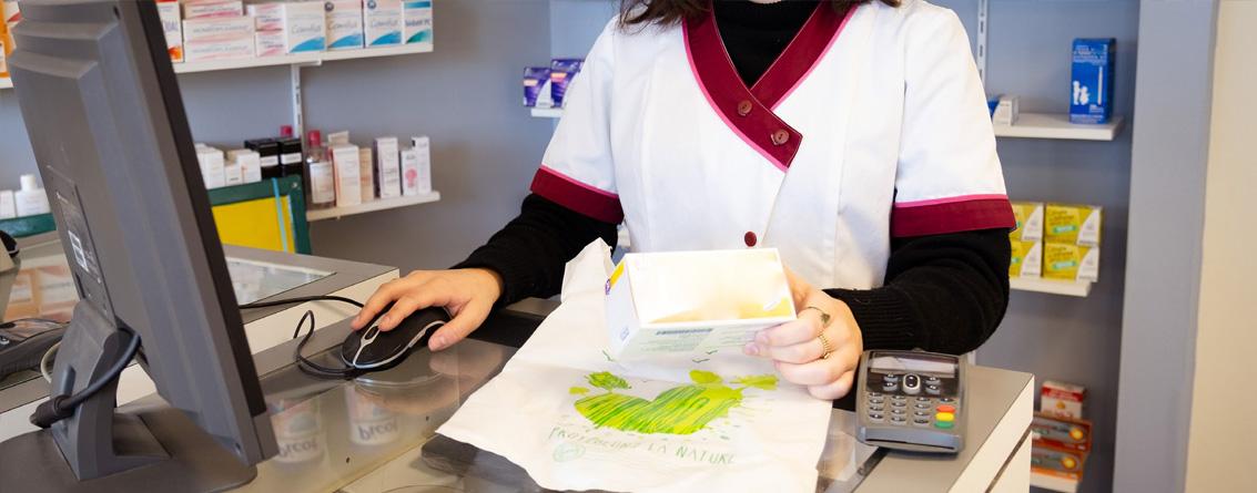 Une jeune préparatrice en pharmacie avec une boite de médicament à la main regarde son écran d'ordinateur