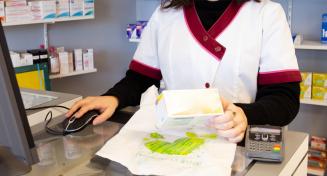 Une jeune préparatrice en pharmacie avec une boite de médicament à la main regarde son écran d'ordinateur